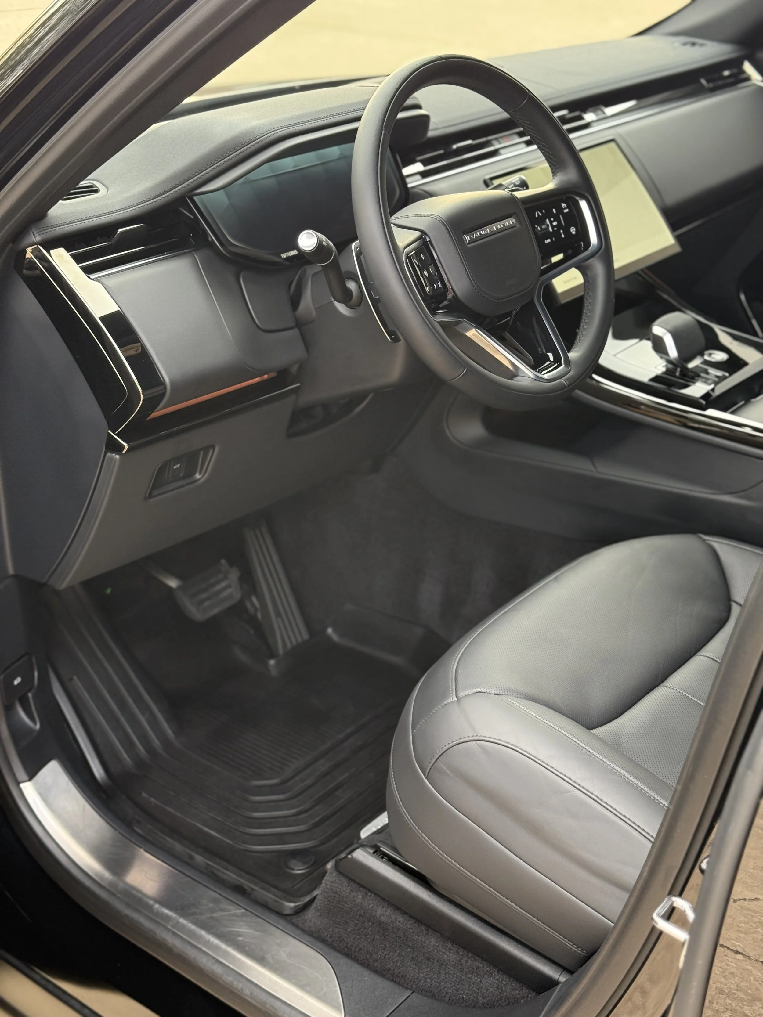Interior of a modern vehicle showing a leather seat, a steering wheel with various controls, a digital dashboard, and a clean center console with a gear shifter and touch screen display.
