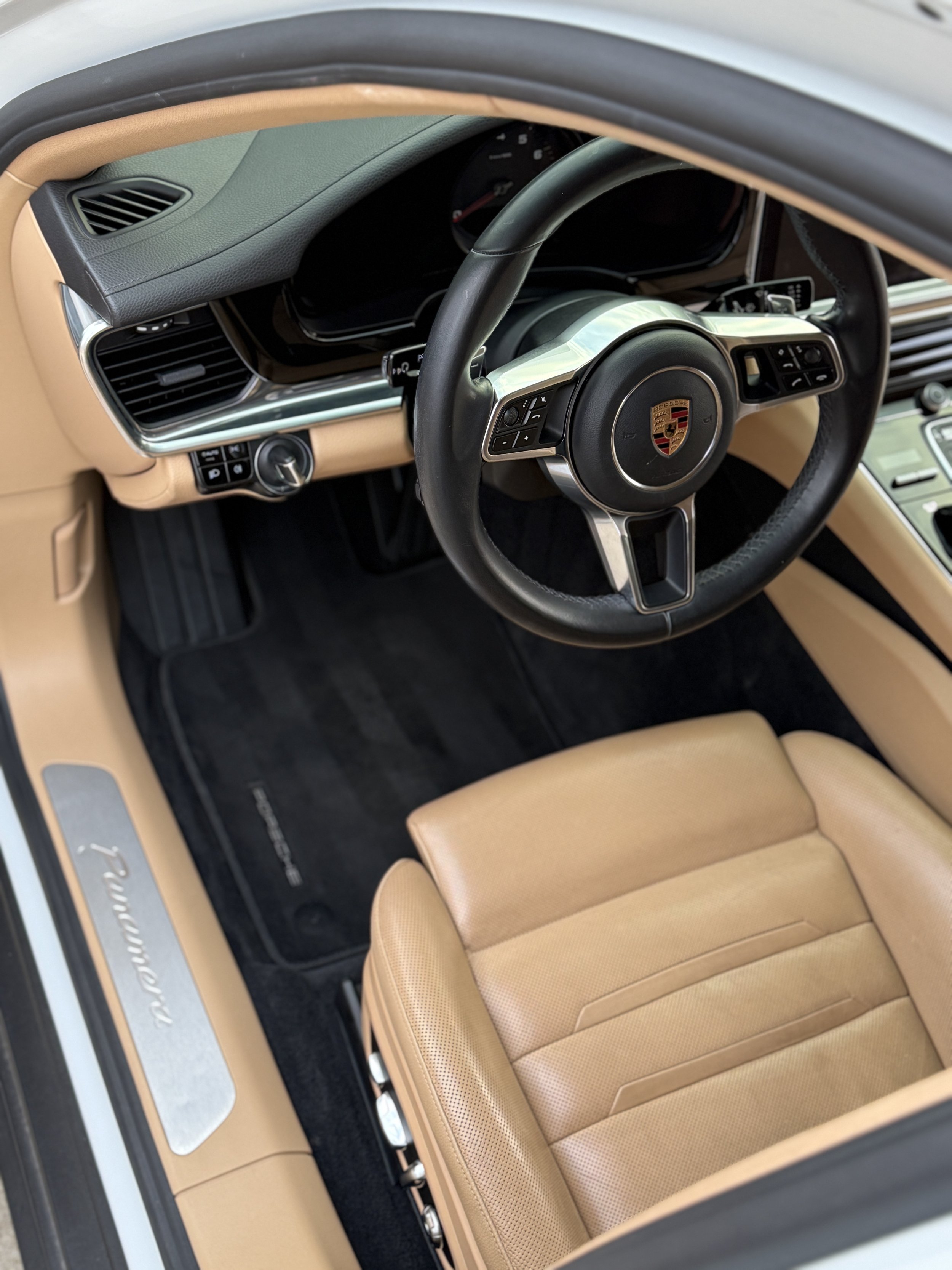 The interior of a beige Porsche vehicle showing the steering wheel with the Porsche logo, tan leather seat, black floor mat, and dashboard with controls and air vents.