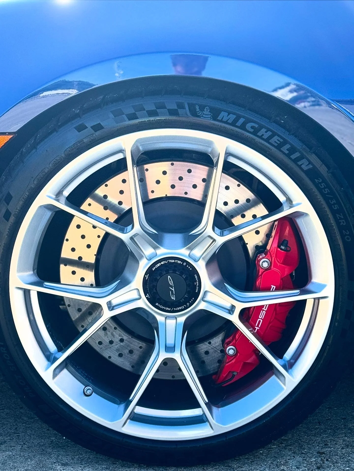 Close-up of a car wheel with a silver rim, black tire, ventilated brake disc, and a red brake caliper, with the car's blue body visible at the top.