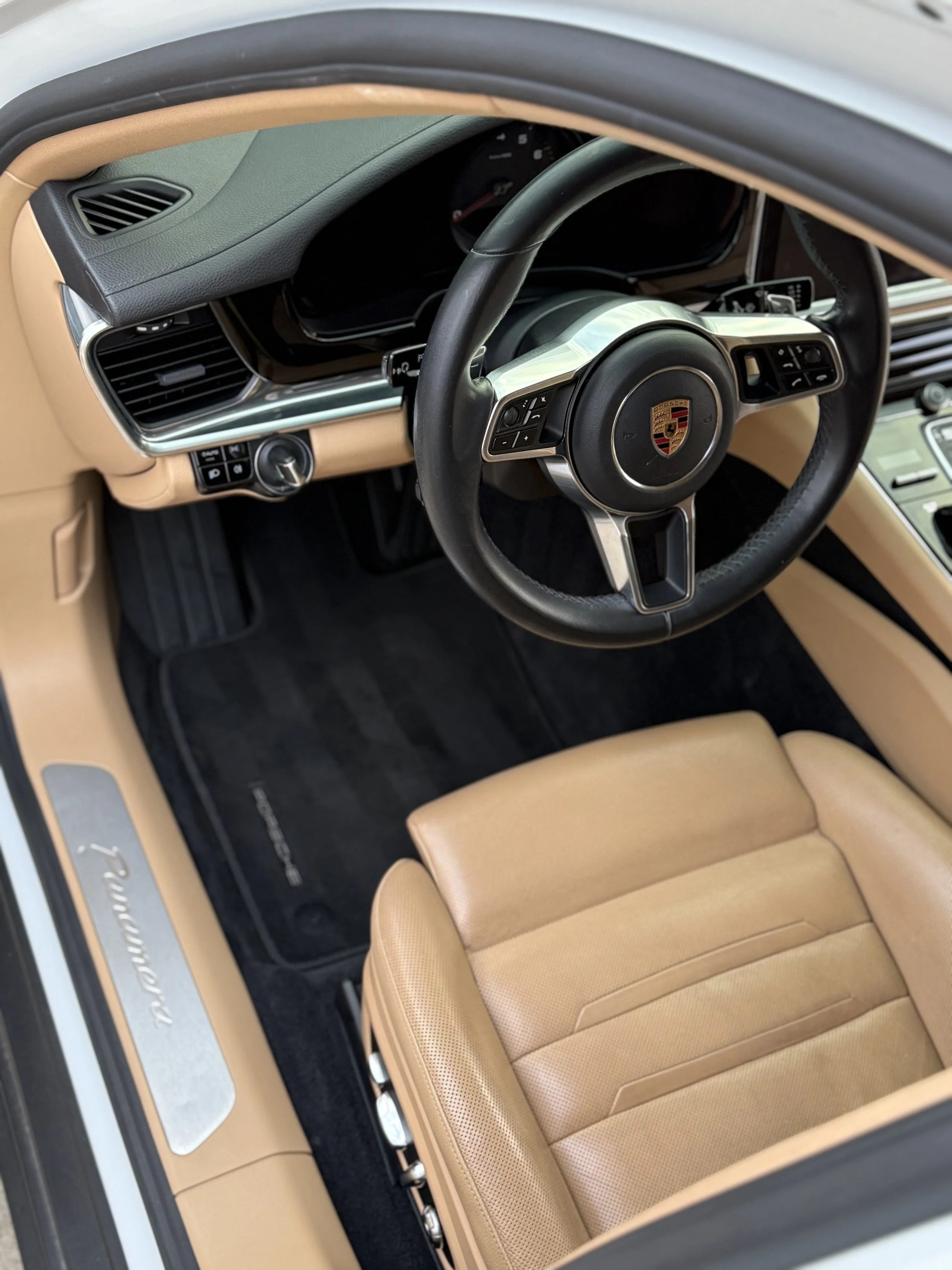 Interior of a Porsche vehicle featuring a tan leather seat, black steering wheel with Porsche crest, and beige dashboard with black trim.