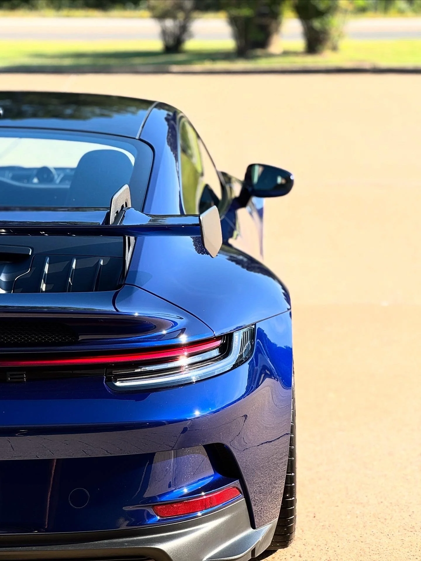 Close-up of the front and side of a shiny blue sports car with a rear spoiler, parked outdoors on a sunny day with trees and grass in the background.