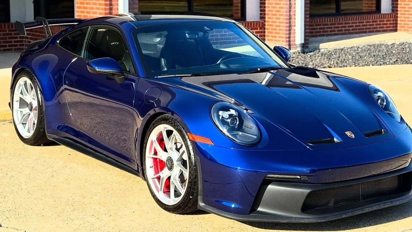 A blue Porsche 911 sports car with white wheels and red brake calipers, parked in front of a brick building on a yellow concrete surface.