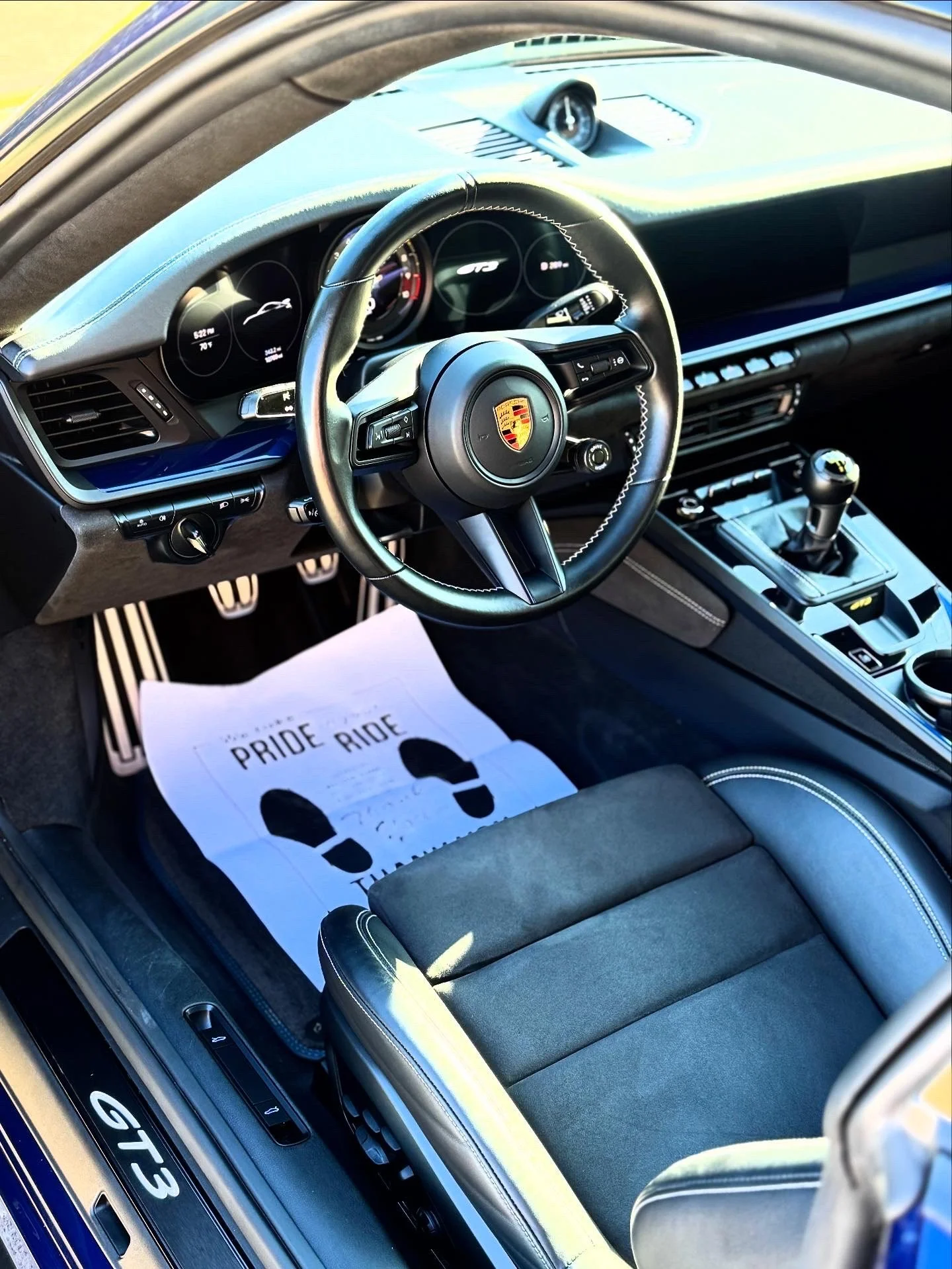 Interior of a Porsche GT3 sports car showing steering wheel, dashboard, gear shifter, and a paper floor mat with the words "PRIDE RIDE".