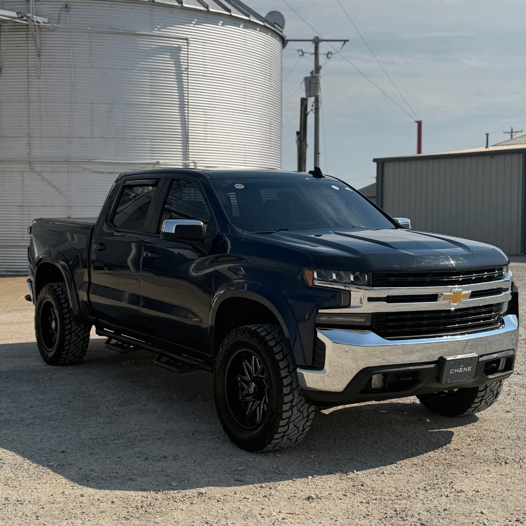 Black Chevrolet pickup truck parked on dirt in front of a large silos and a metal building with power lines in the background.