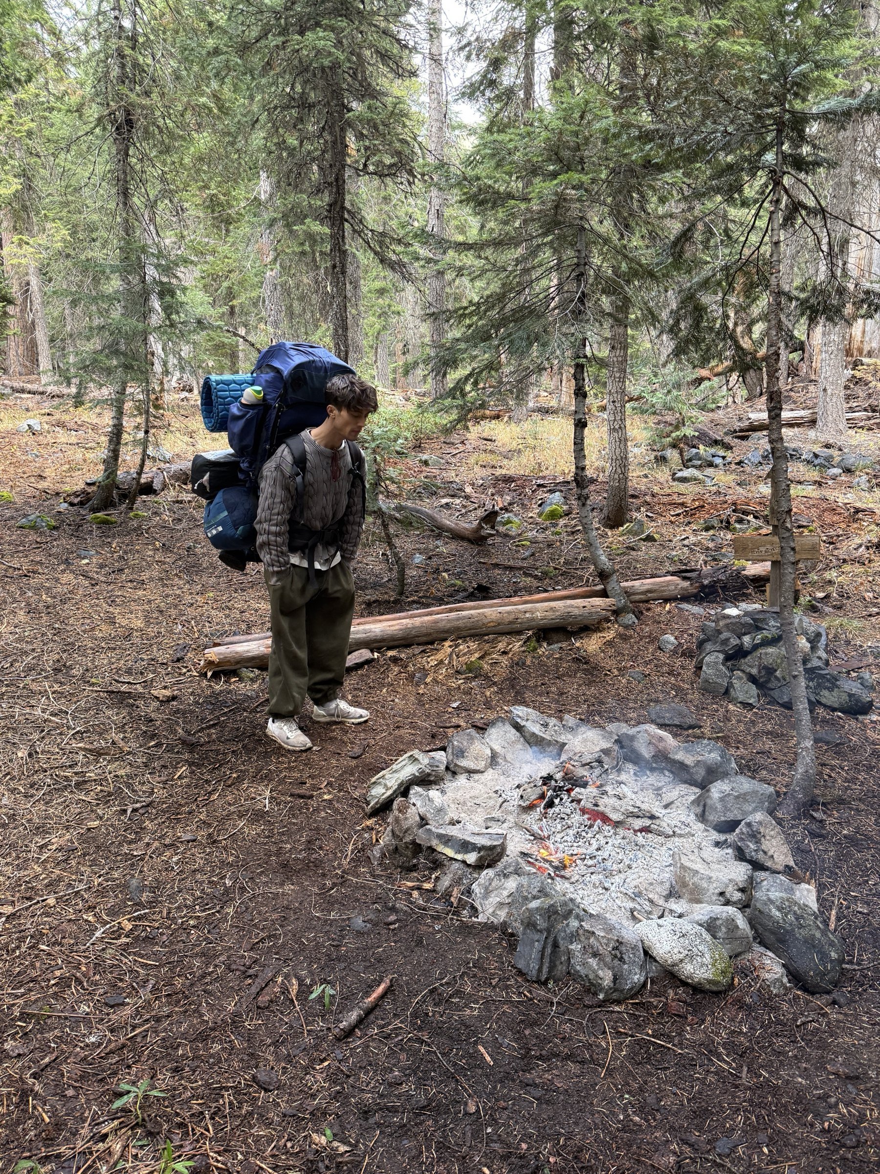 Eli standing with his pack beside the fire ring at camp, with Kevin's sign and a small cairn behind him