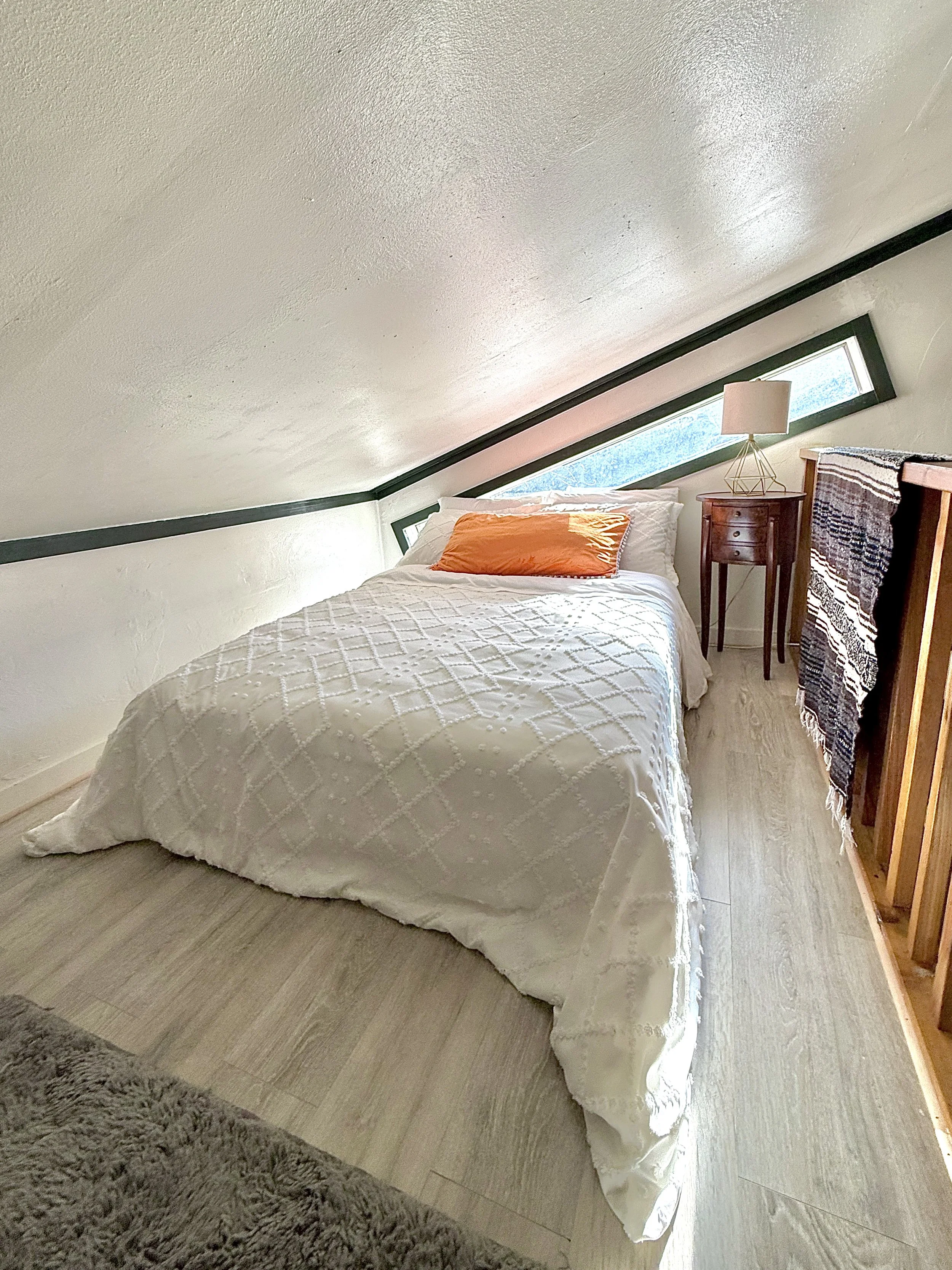 A cozy bedroom with a slanted ceiling, a bed with white bedding and an orange pillow, a small wooden nightstand with a lamp, and a window with natural light.