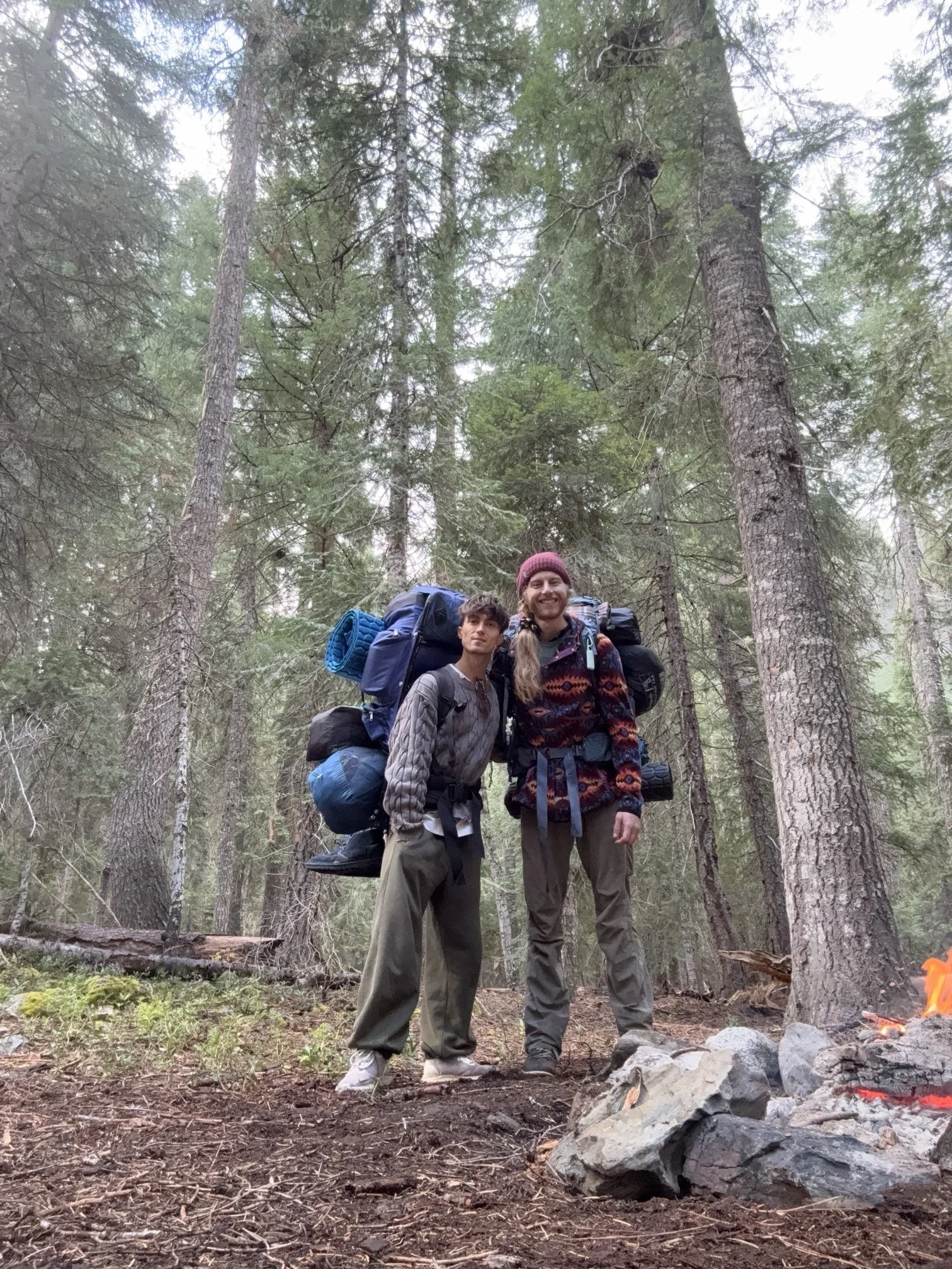 Eli and Colt side by side with full packs under tall firs, ready to hike out