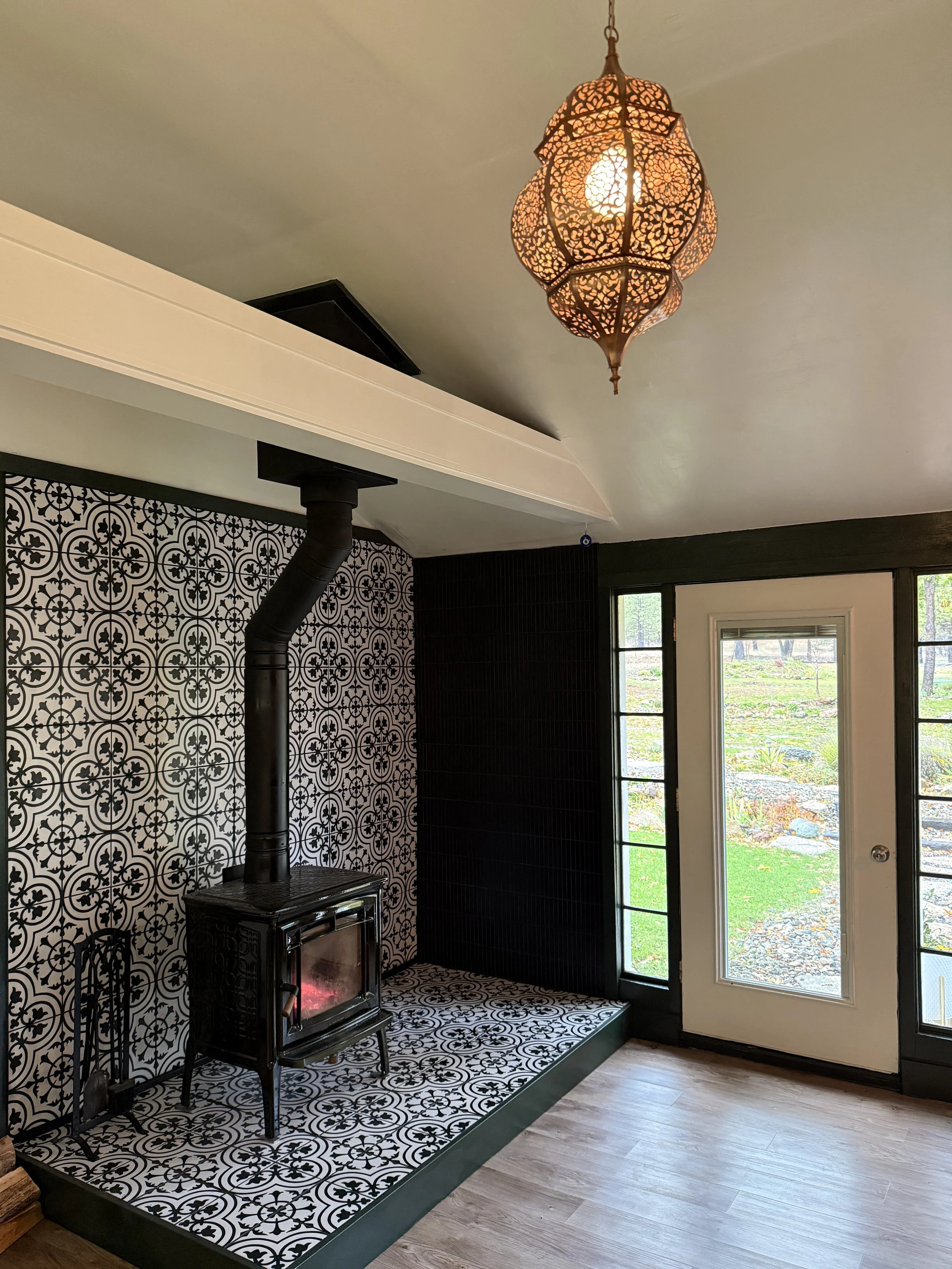 Living room with black and white patterned tiles, a black wood stove with a pipe, large windows, a door leading outside, and a Moroccan-style hanging lamp.