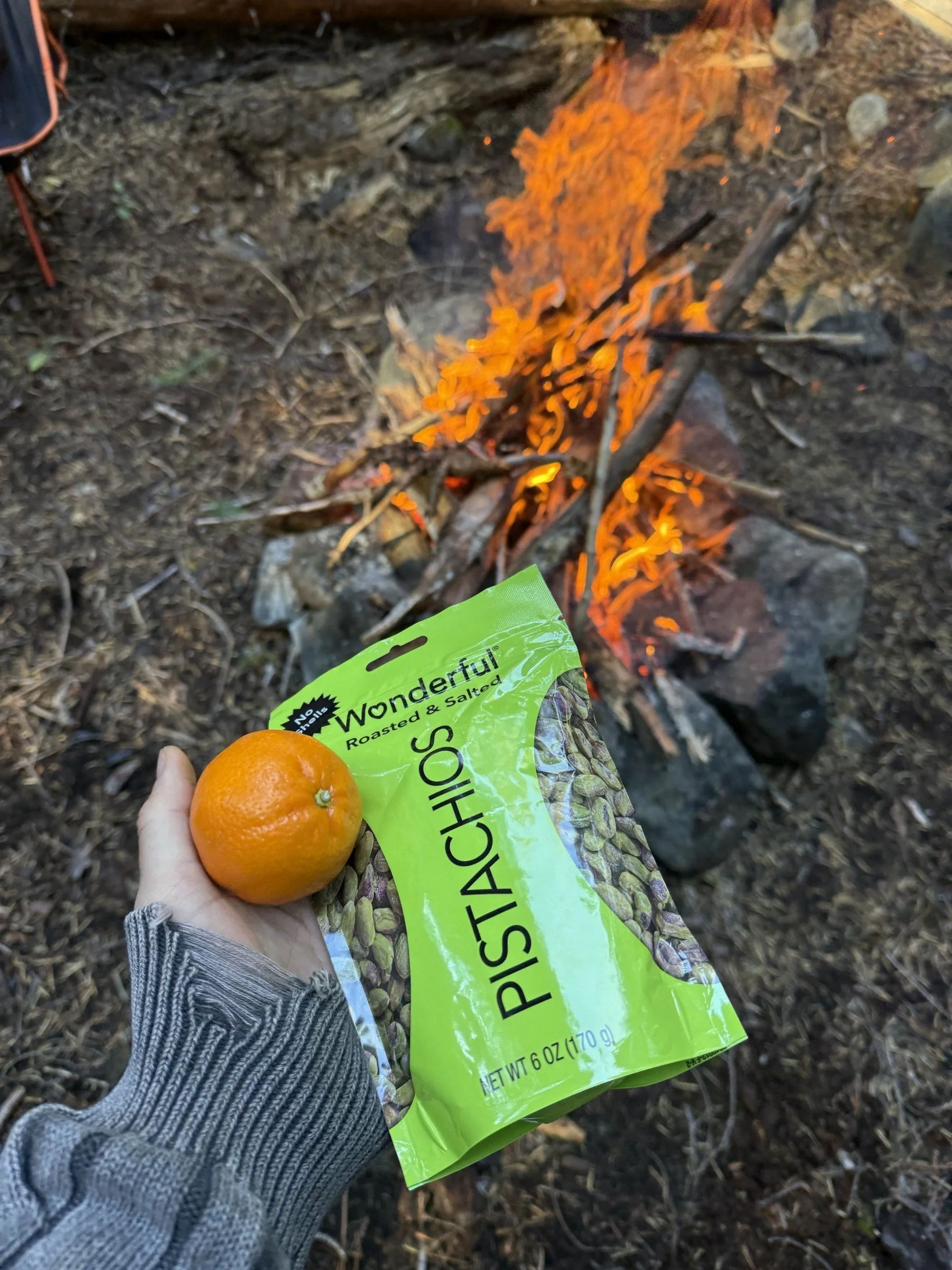 An orange and a bag of pistachios held up in front of the campfire