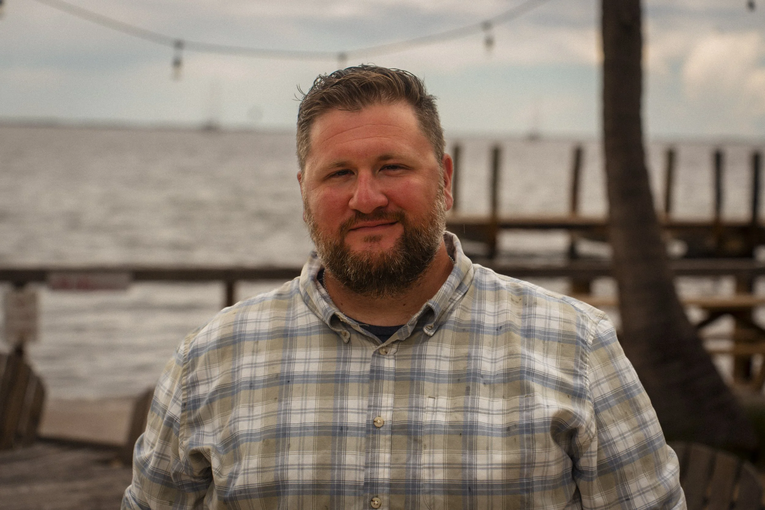 A young man with a beard and light hair in a button up shirt, standing outdoors with a blurred ocean background.