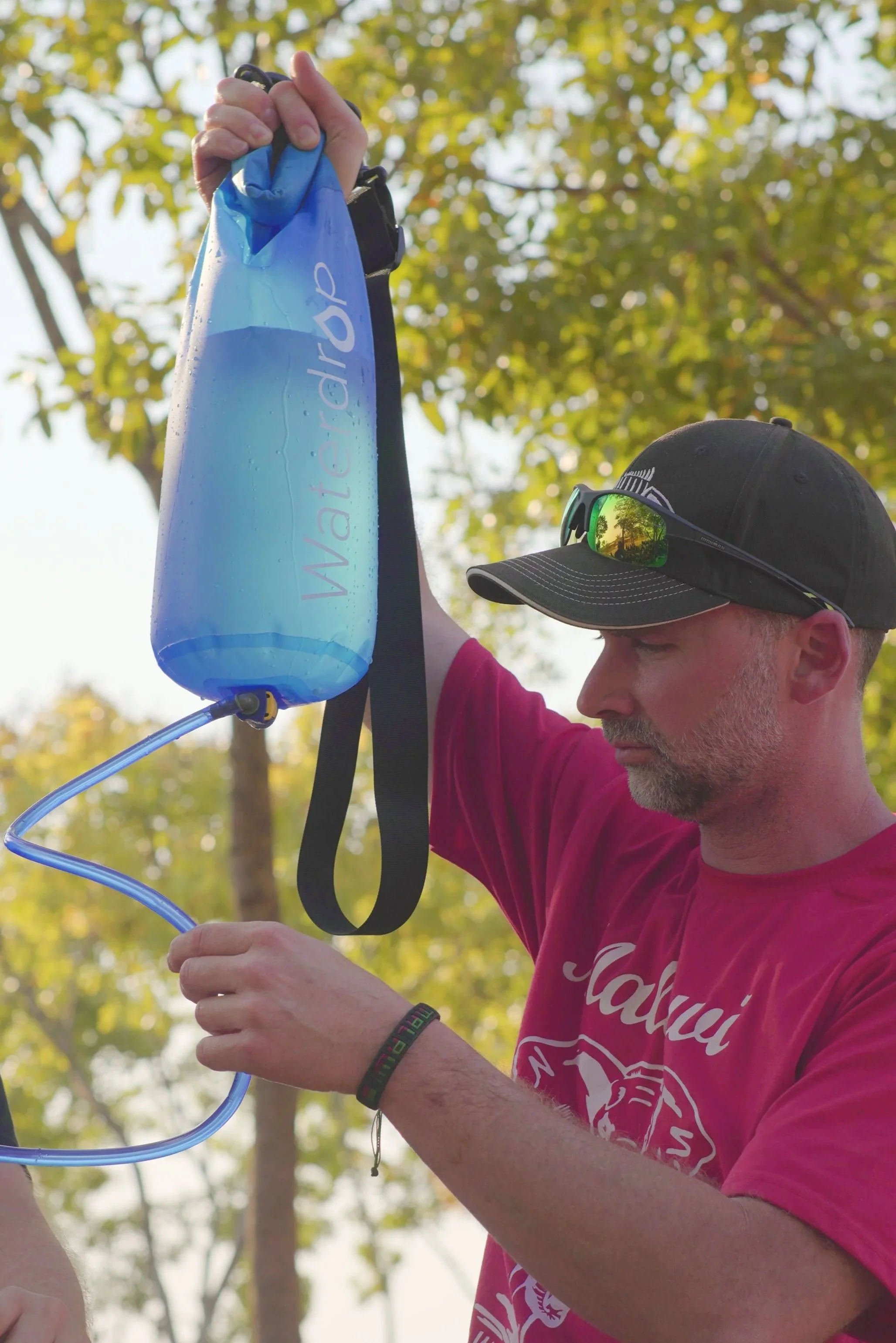 A man wearing a black cap and sunglasses on his head is filling a blue water bladder from a hydration tube outdoors in a wooded area.