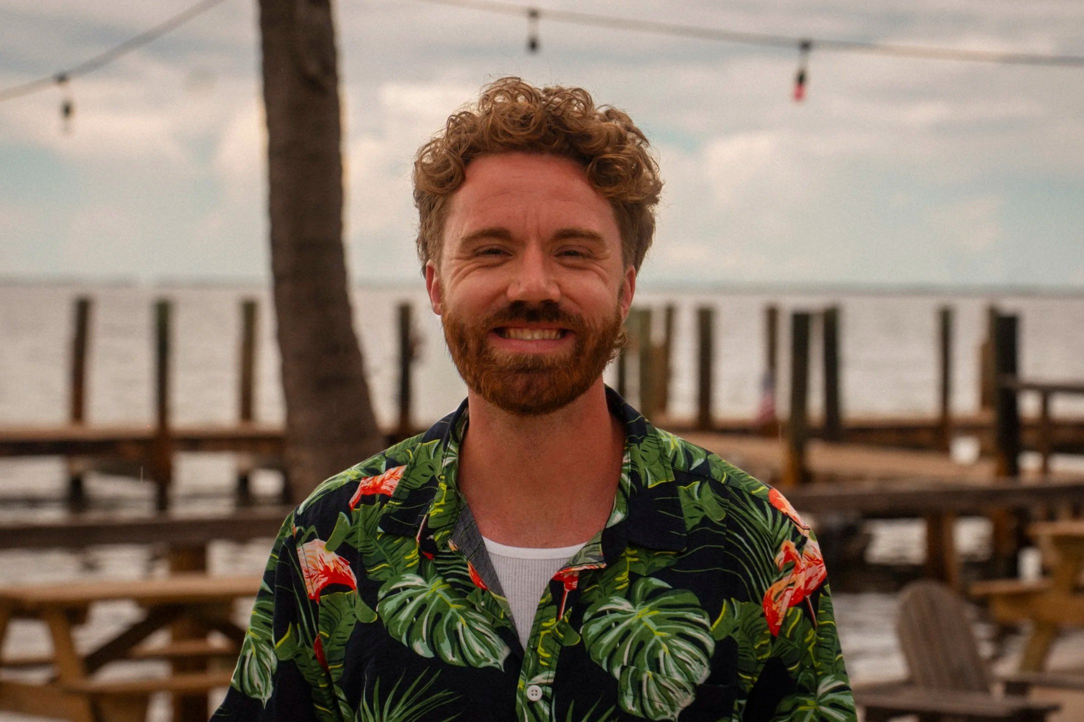 Portrait of a smiling man with a beard and short wavy hair, wearing a plaid shirt, outdoors with blurred green foliage background.