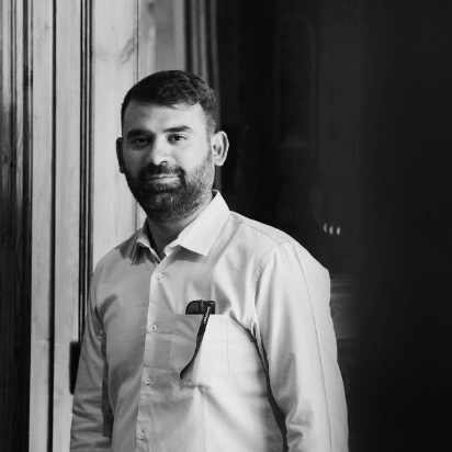 Black and white photo of a man with a beard, wearing a button-up shirt, standing indoors near wooden paneling, with a pen in his shirt pocket.