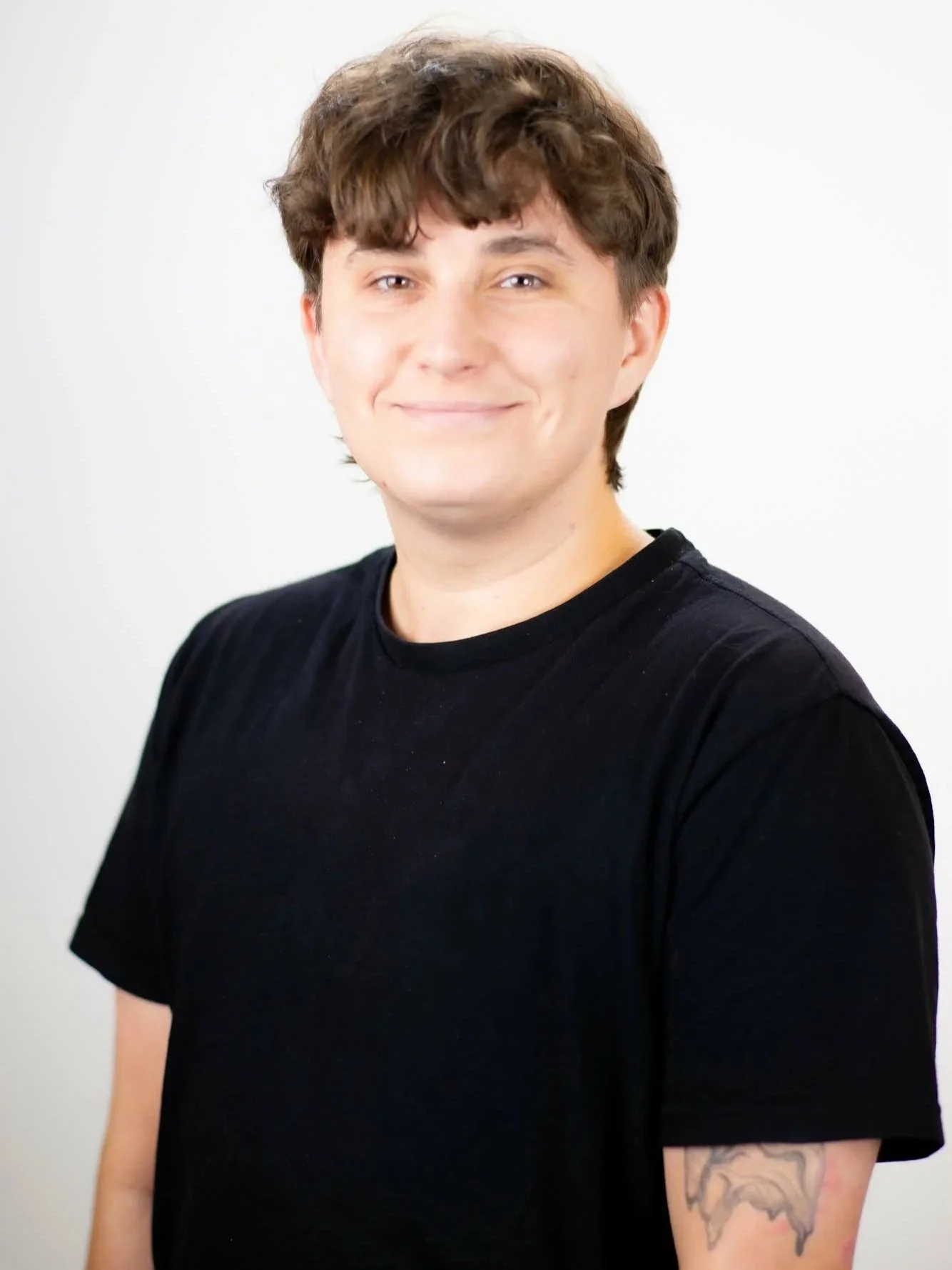 Young trans nonbinary poet with short brown curly hair, wearing a black T-shirt, smiling, with a tattoo on their upper arm, standing against a plain white background.