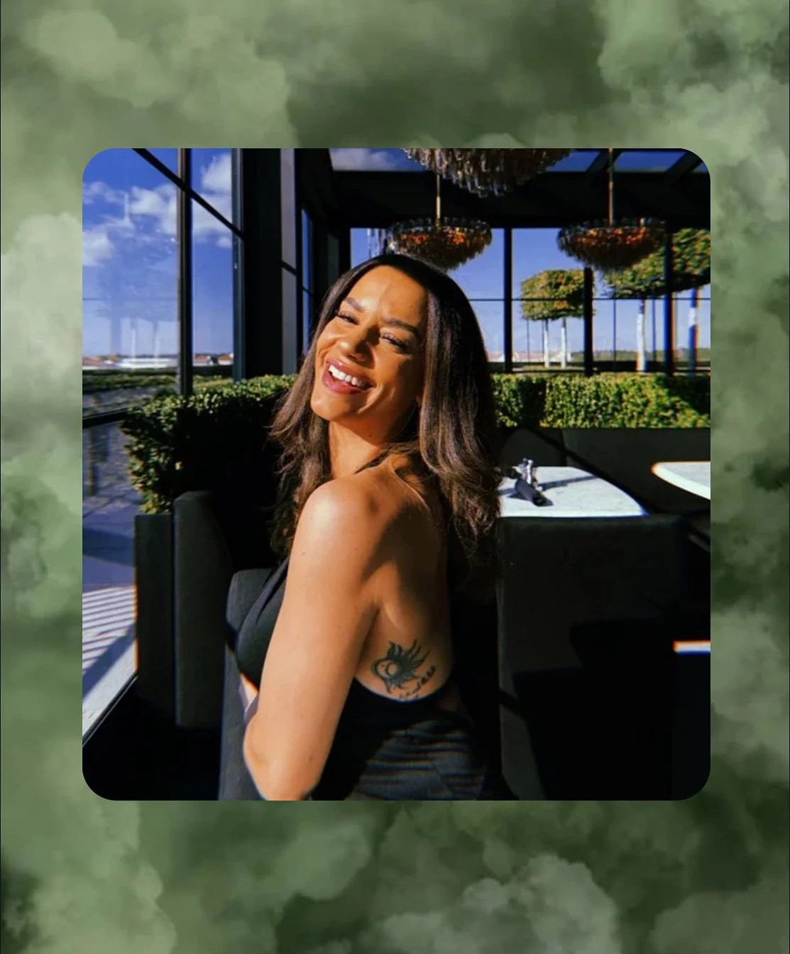 Smiling woman with dark hair and tattoo on her upper arm, sitting in a bright restaurant with large windows, greenery outside, and chandeliers hanging from the ceiling.