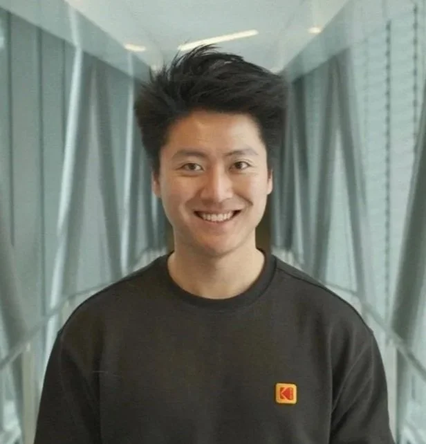 A smiling young man with black hair standing in an indoor hallway with glass walls and curtains.