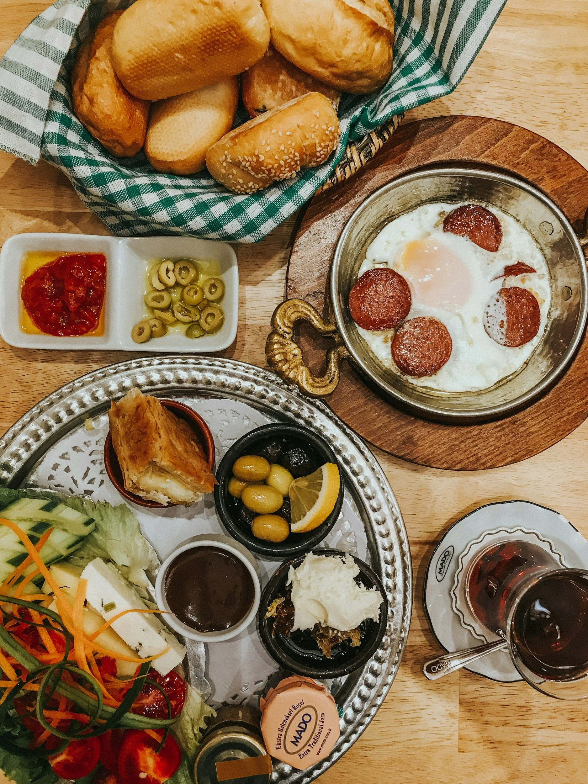 A breakfast spread featuring a basket of mixed bread rolls, a skillet with fried eggs and slices of sausage, a tray with salad, olives, a lemon wedge, and small dishes of jam, honey, and preserves, plus a glass of red wine.