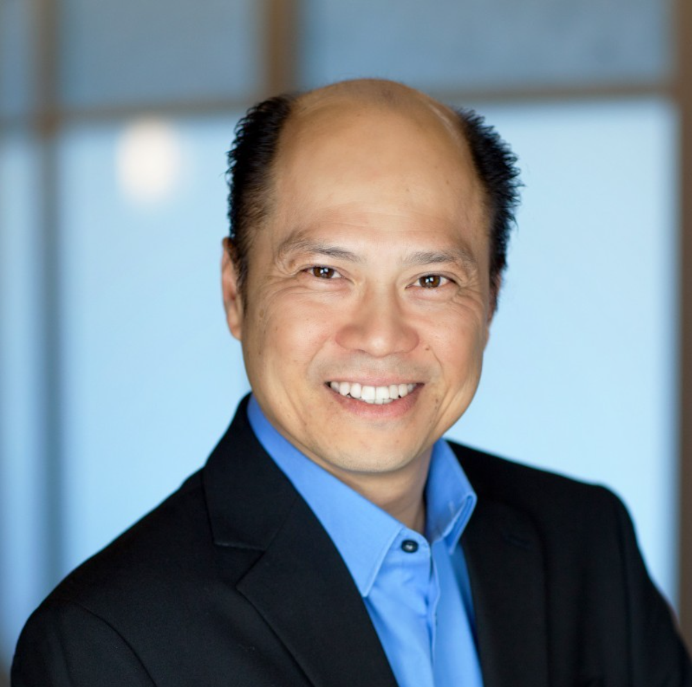 Close-up portrait of a smiling man with a shaved head, wearing a blue shirt and black blazer, standing in front of a blurred indoor background.