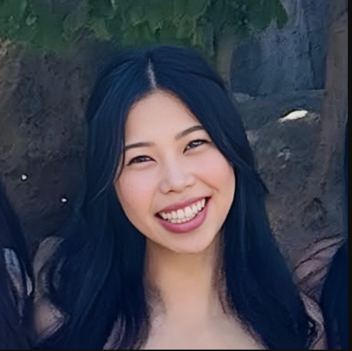 A young woman with long, dark blue hair smiling outdoors in front of a tree.