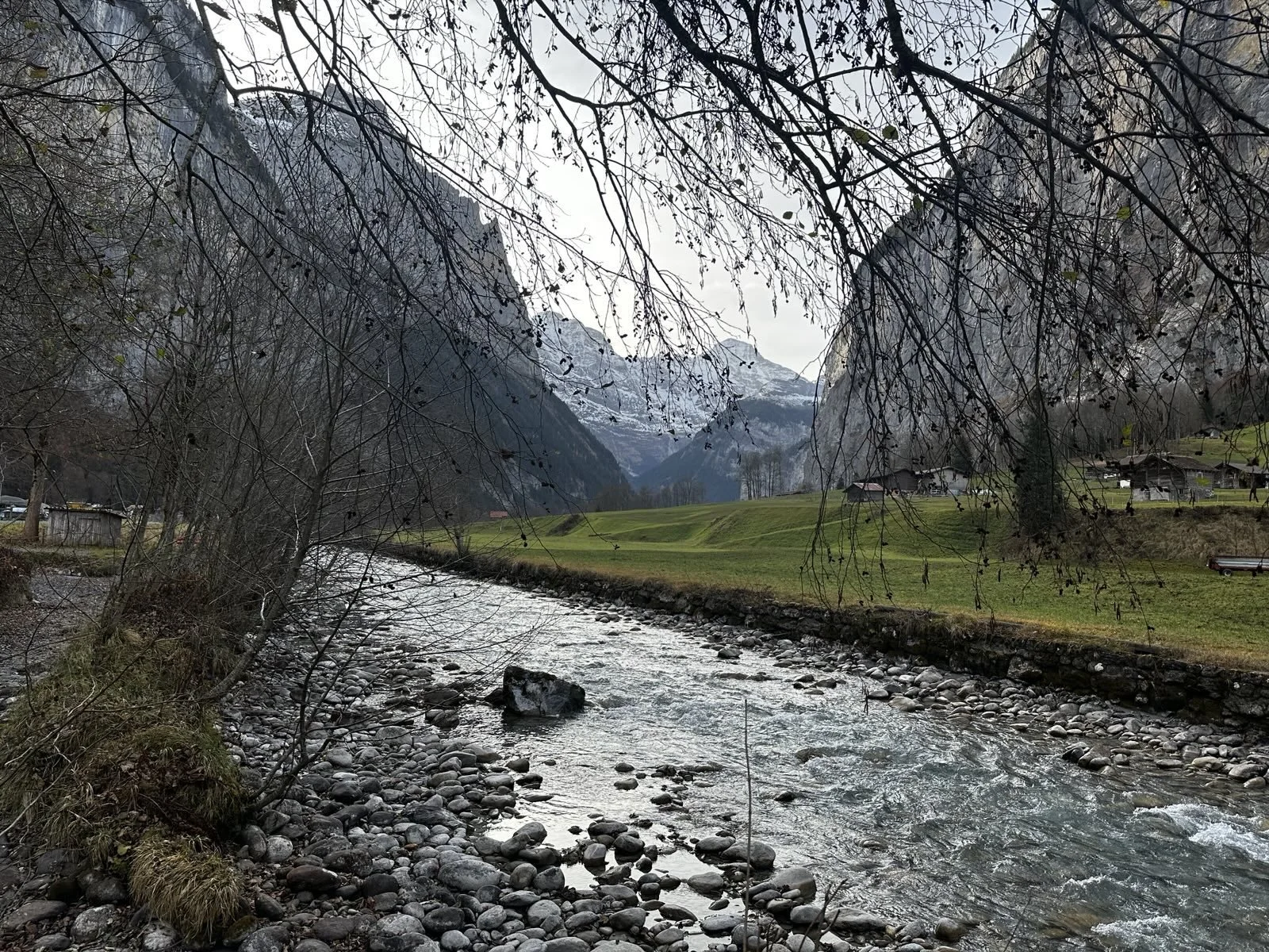 A river flowing through a valley with mountains in the background, trees with bare branches in the foreground, and a green grassy area with small houses on the right side.