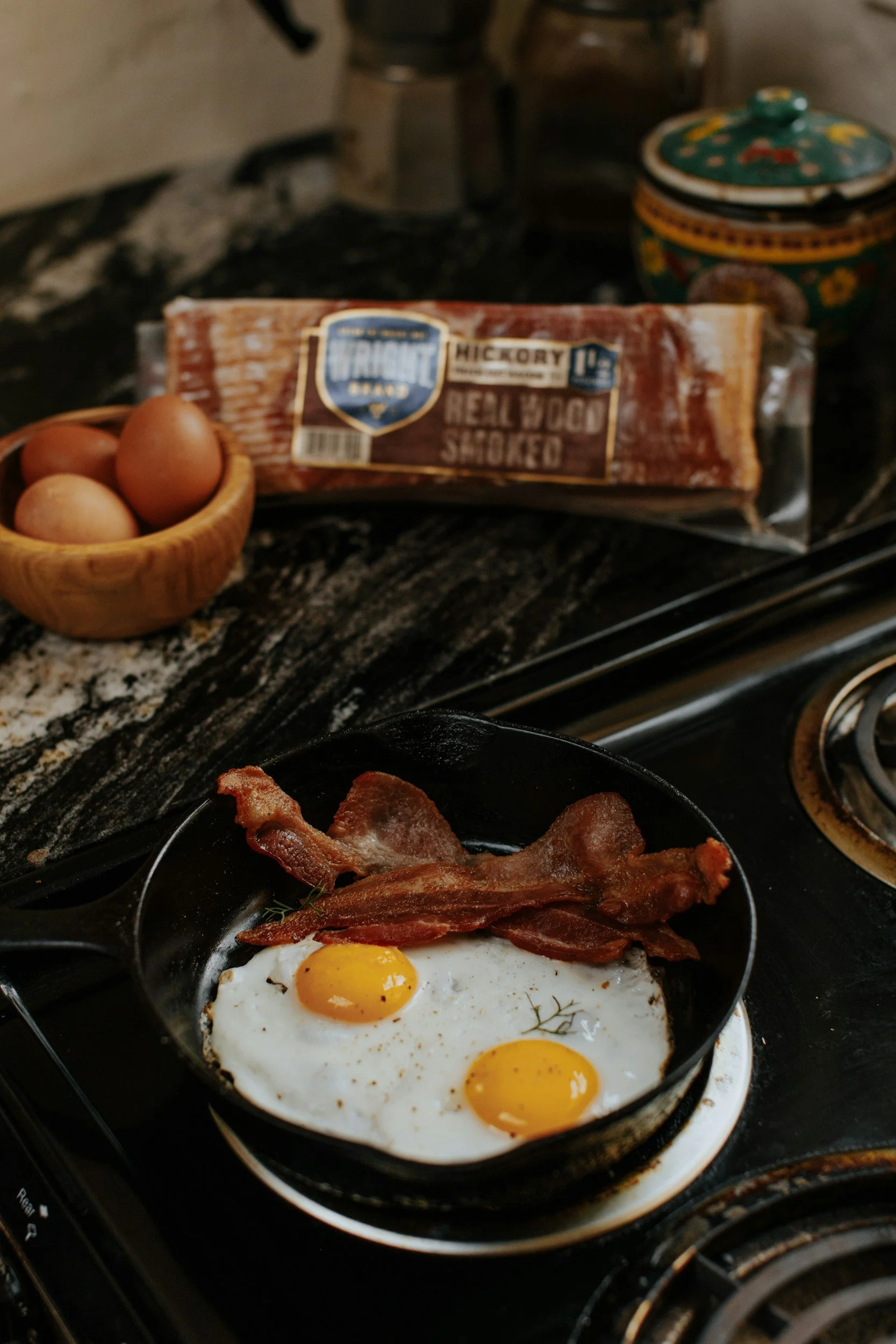 Fried eggs with bacon on a cast iron skillet, background shows eggs and packaged smoked bacon, stove top with a decorative jar.