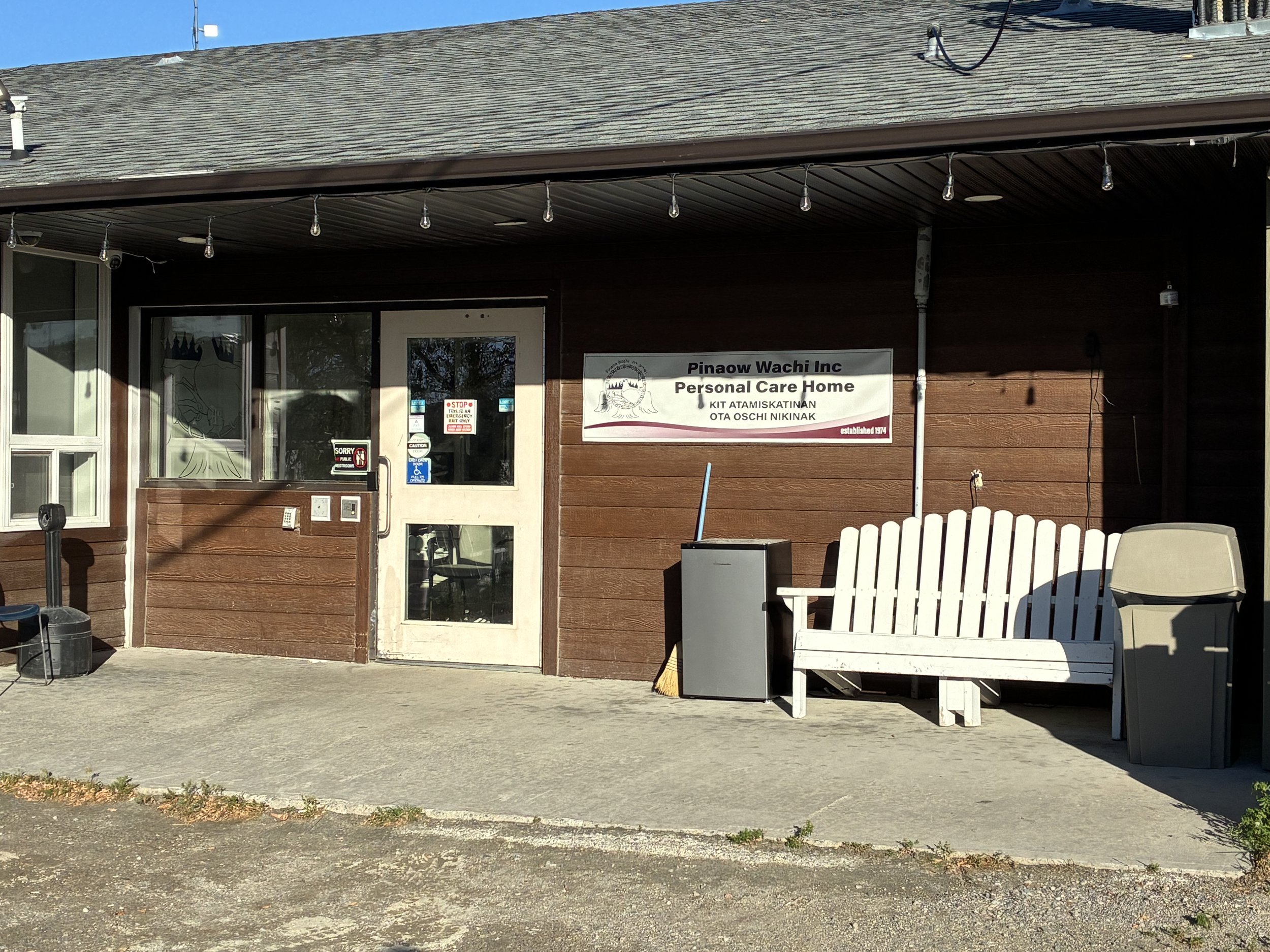The front of a community care home building with a sign reading 'Pinaow Wachi Inc Personal Care Home' and the establishment year 1974. There is an outdoor area with a white bench, a trash can, and string lights hanging under the roof.