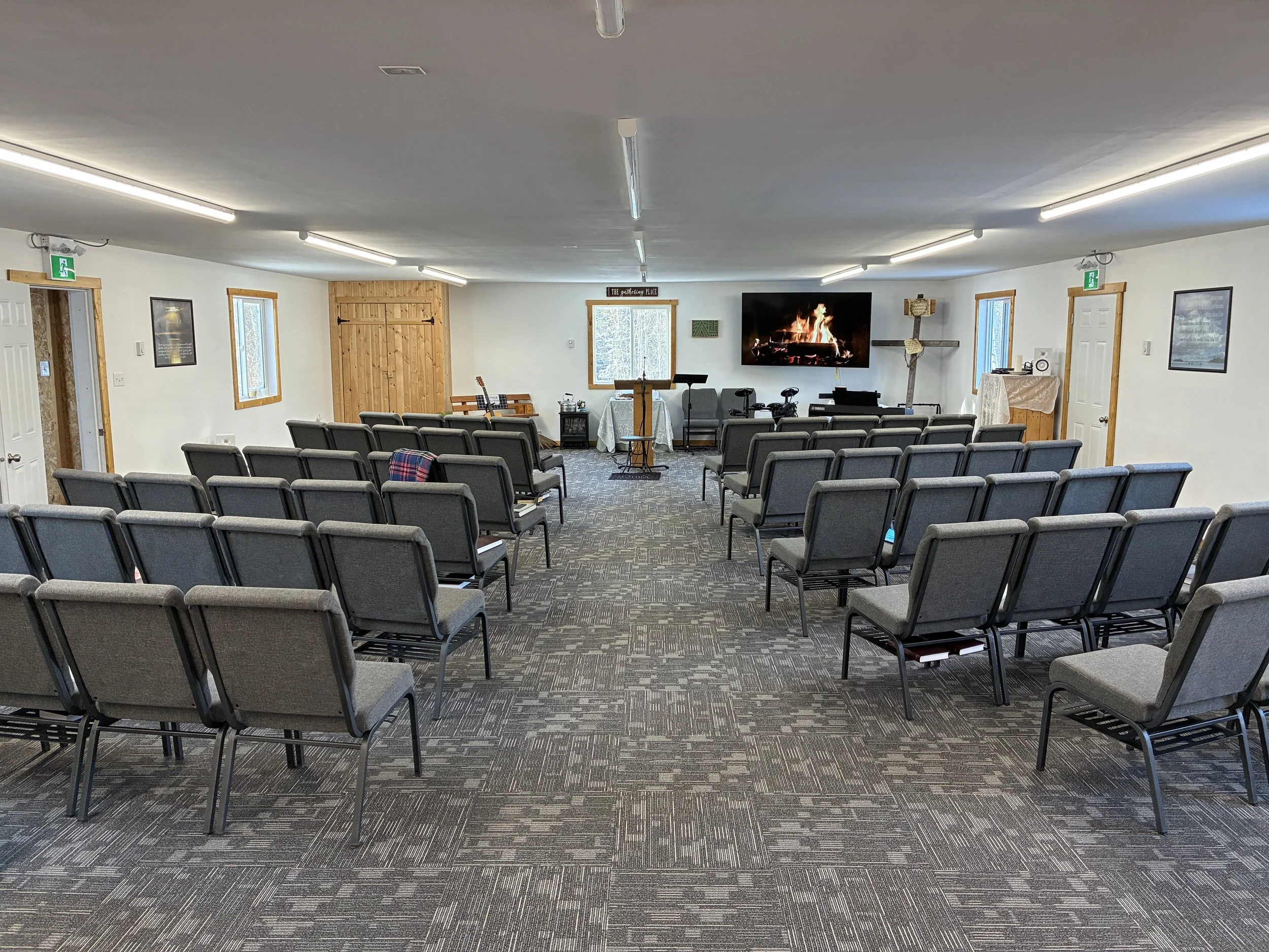 Indoor meeting space with rows of gray chairs facing a large television screen displaying a fireplace scene, with a wooden door and windows on the walls, and a table with a white cloth and a speaker at the front.