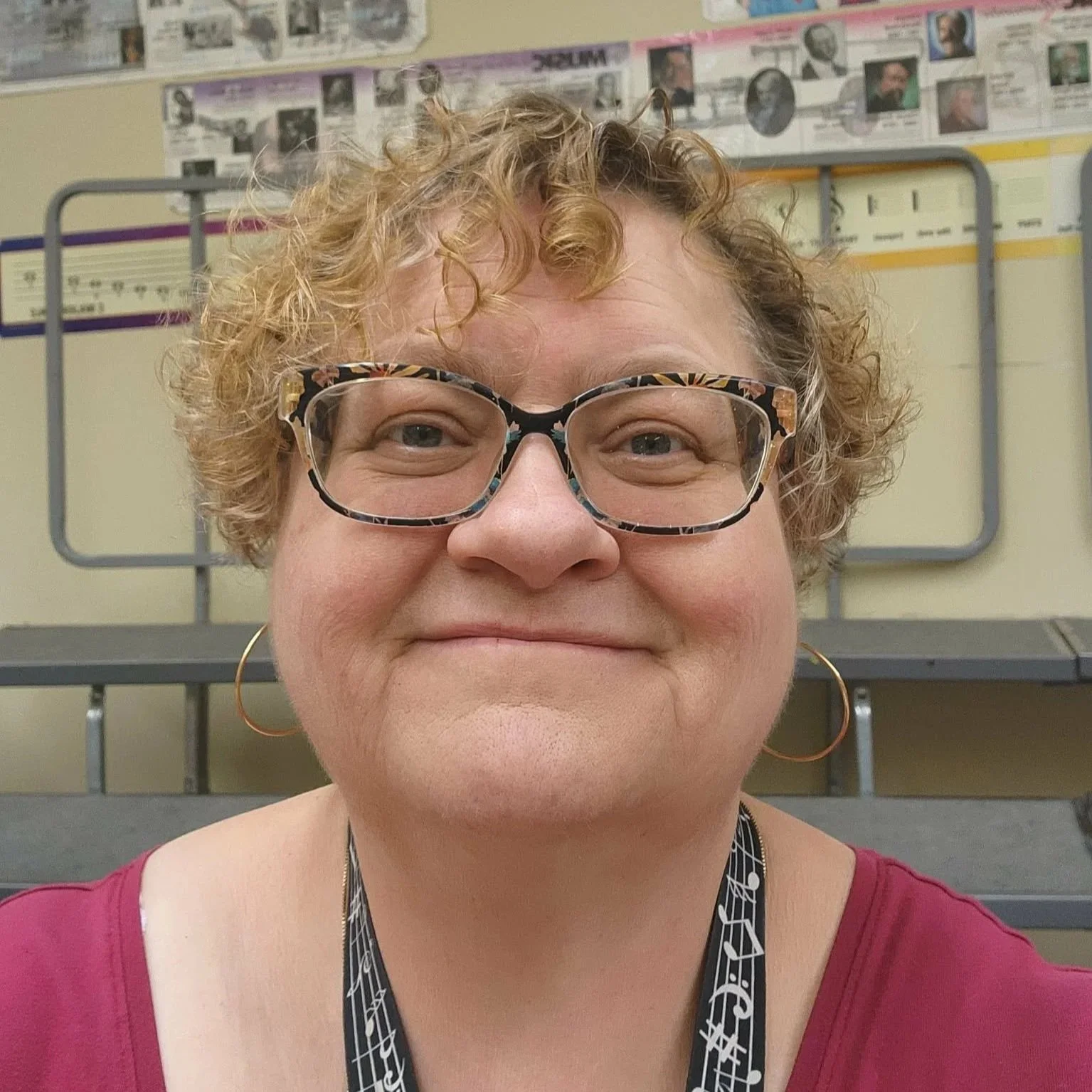 Close-up of a smiling woman with curly hair, wearing patterned glasses, gold hoop earrings, and a music-themed lanyard, seated in a classroom or auditorium with bulletin boards and benches in the background.