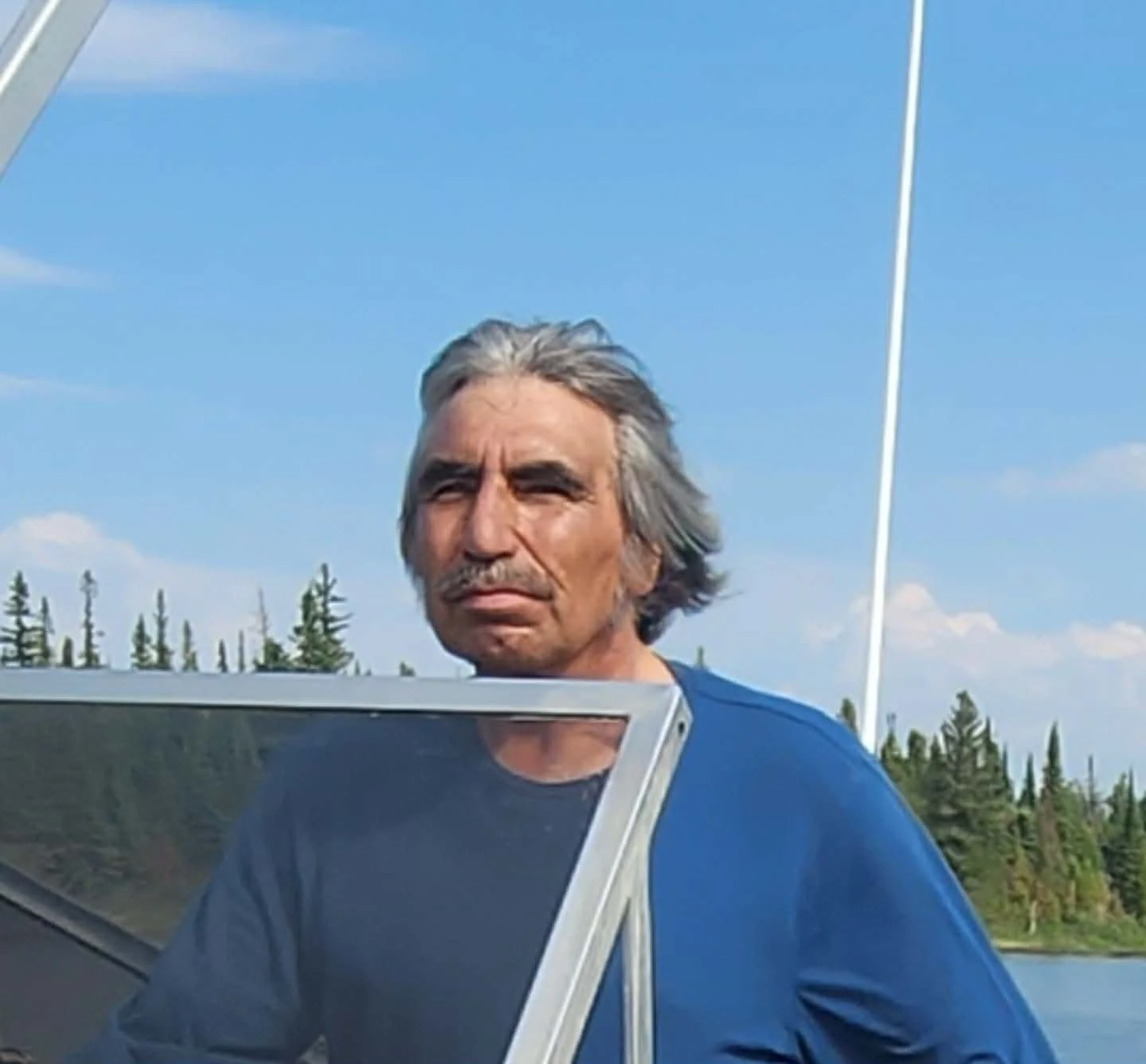 A middle-aged man with long gray hair, a mustache, and wearing a blue shirt, standing outdoors near a body of water with a forest in the background, under a blue sky with some clouds.