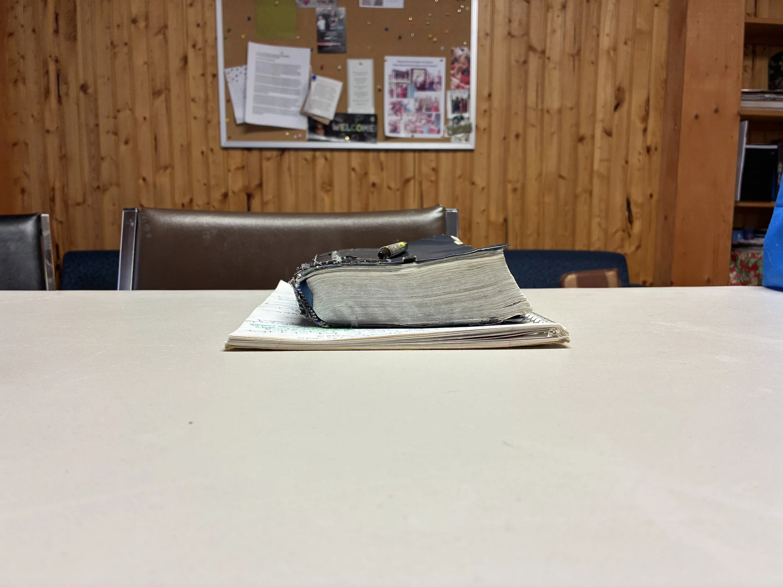 A worn, damaged Bible resting on open notebooks on a white table in a wooden-paneled room, with a bulletin board and chairs in the background.
