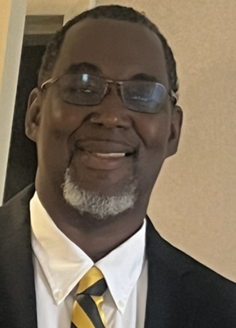 Smiling African American man wearing glasses, a white shirt, a black suit, and a yellow and black striped tie, indoors with a beige background.