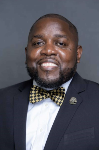 Portrait of a man in a suit with a yellow and black checkered bowtie, smiling against a gray background.