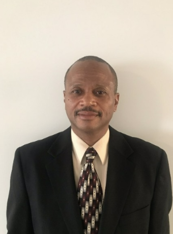 A middle-aged man with a mustache wearing a dark suit, white shirt, and patterned tie, standing against a plain white wall.