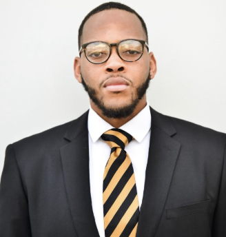 A man wearing glasses, a black suit, white shirt, and a black and yellow striped tie, standing against a plain white background.