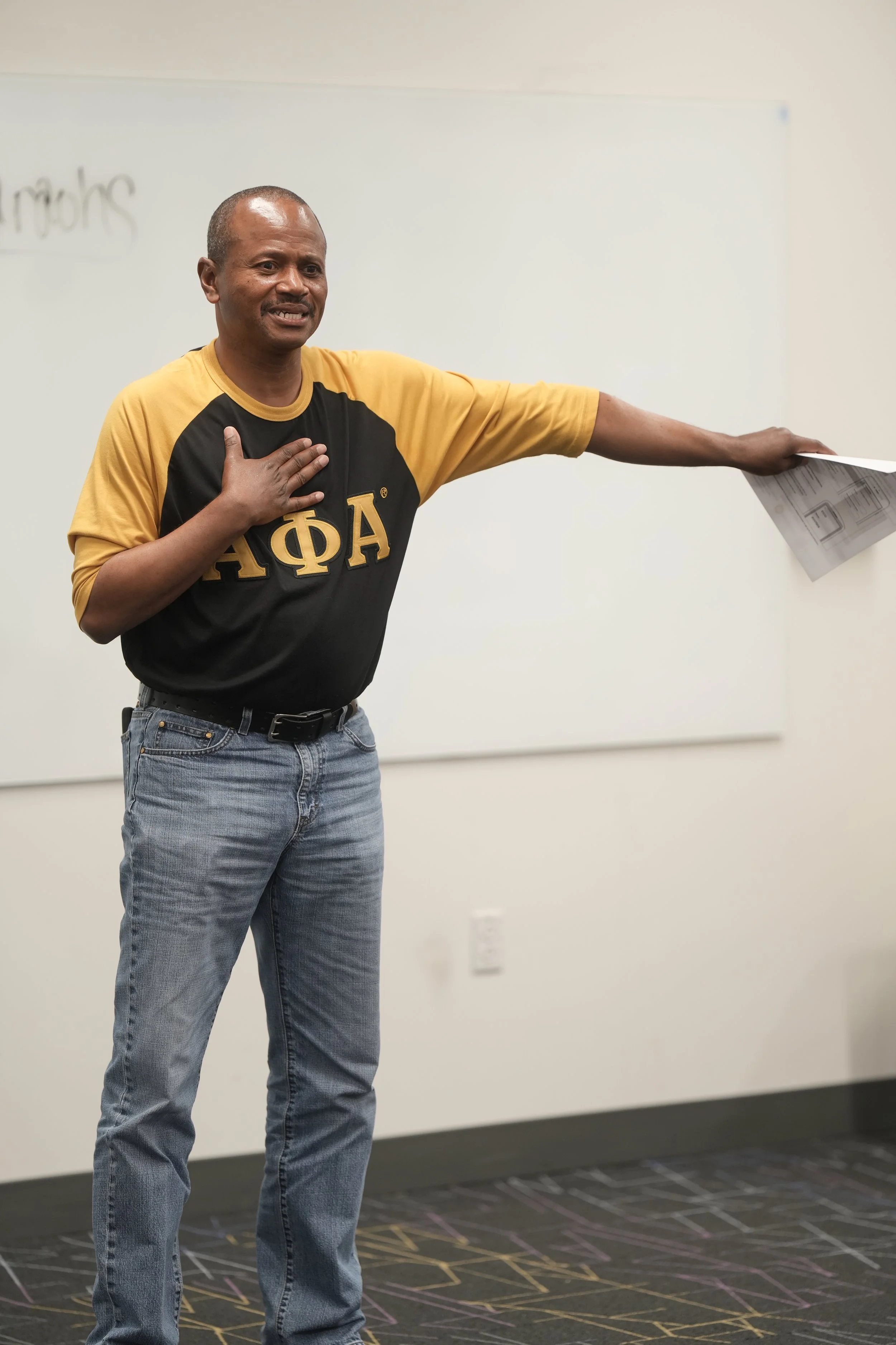 A man wearing a black and gold shirt with Greek letters, standing in front of a whiteboard, with his left arm extended to the side and his right hand over his heart.