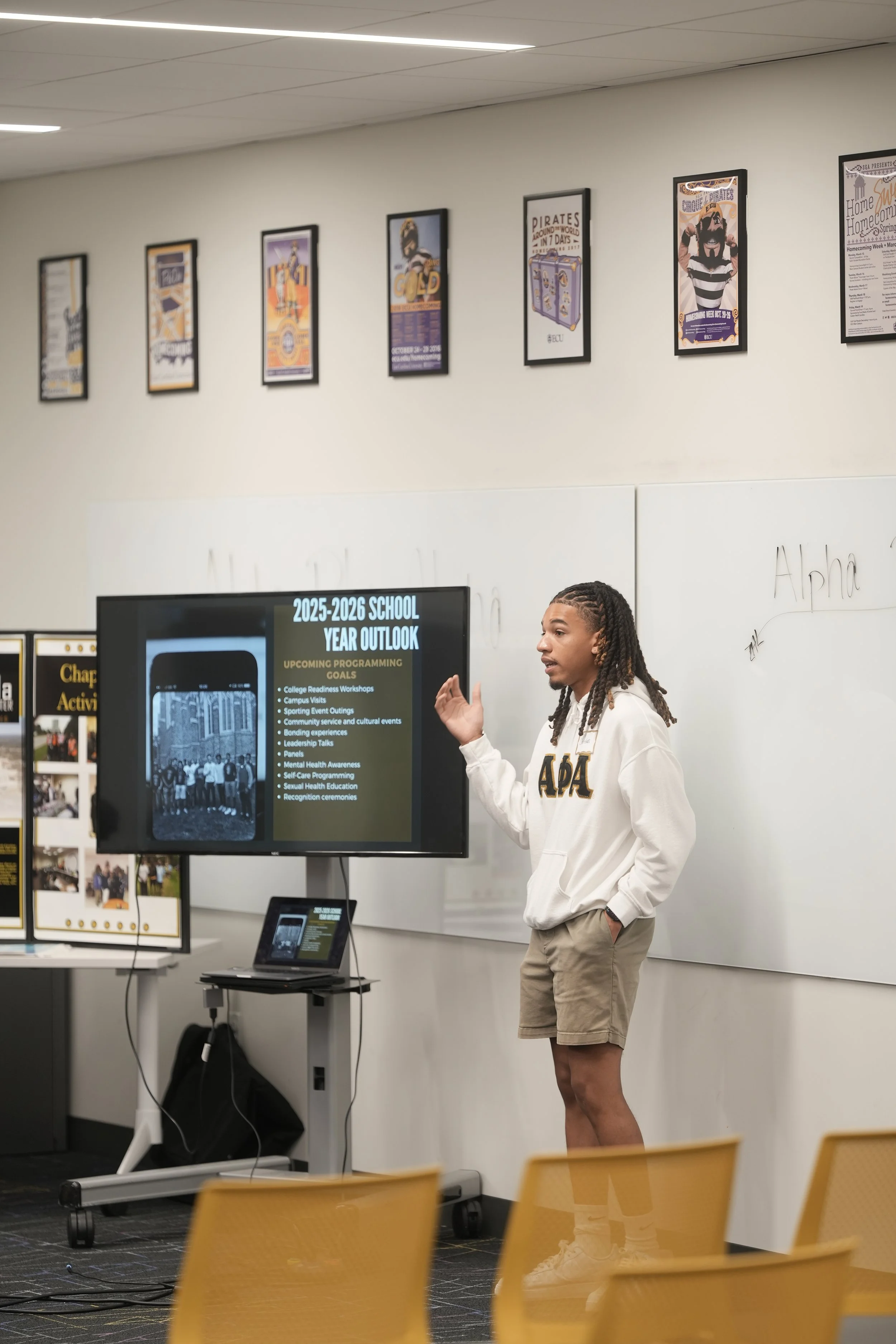 A young woman with braided hair giving a presentation in a classroom. She is standing next to a screen displaying the 2025-2026 school year outlook and upcoming programming goals, wearing a white hoodie with Greek letters and shorts. There are posters on the wall and chairs in the foreground.