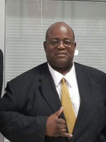Man in a suit and glasses making a shaka sign with his right hand in an office setting.