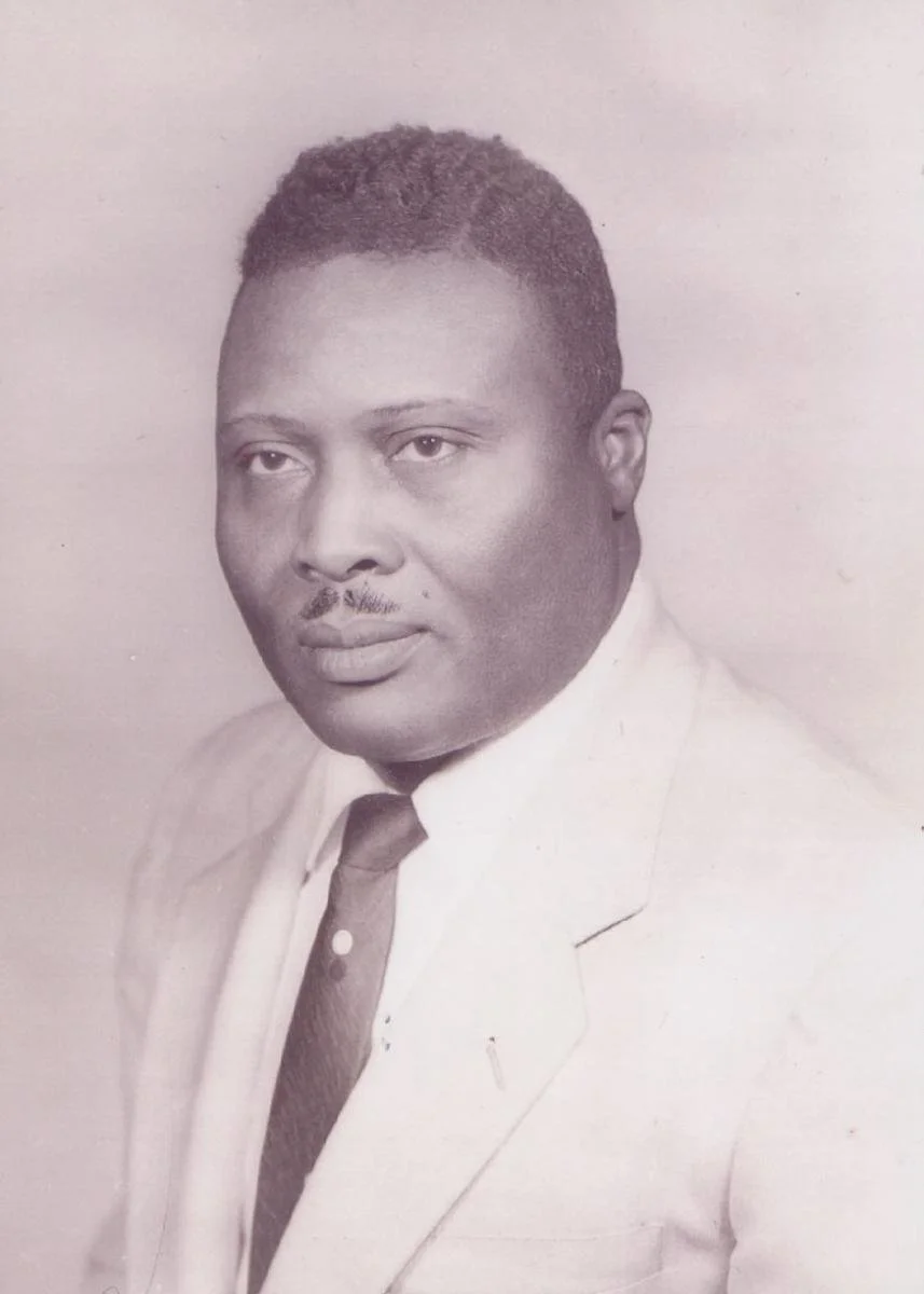 A black-and-white portrait of a man wearing a suit and tie, looking slightly to his right with a serious expression.