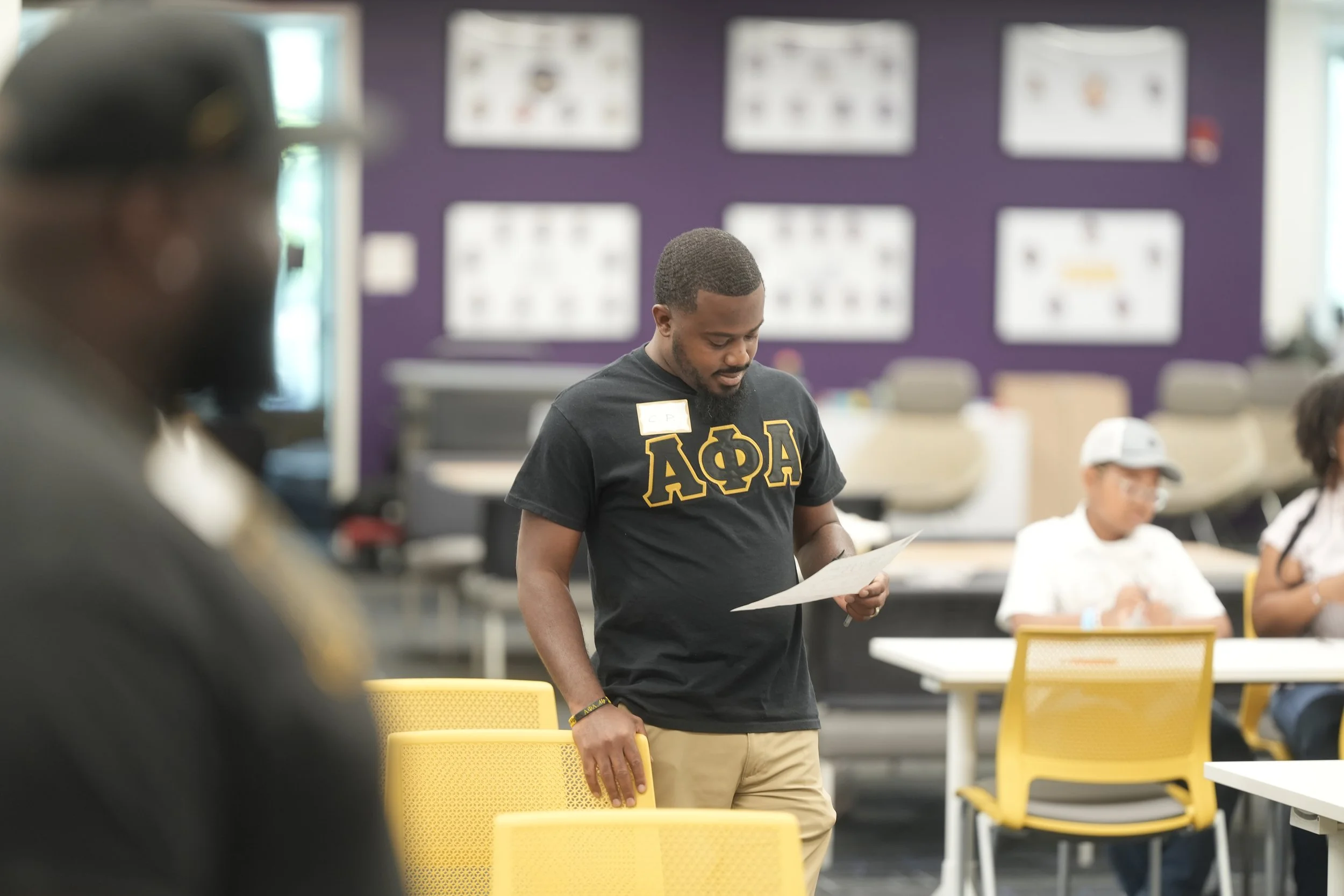 A young man with a beard wearing a black T-shirt with Greek letters and a gold name tag, standing in a classroom or meeting room, reading a paper. Other people are seated at tables around him, and there are posters on the purple wall in the background.