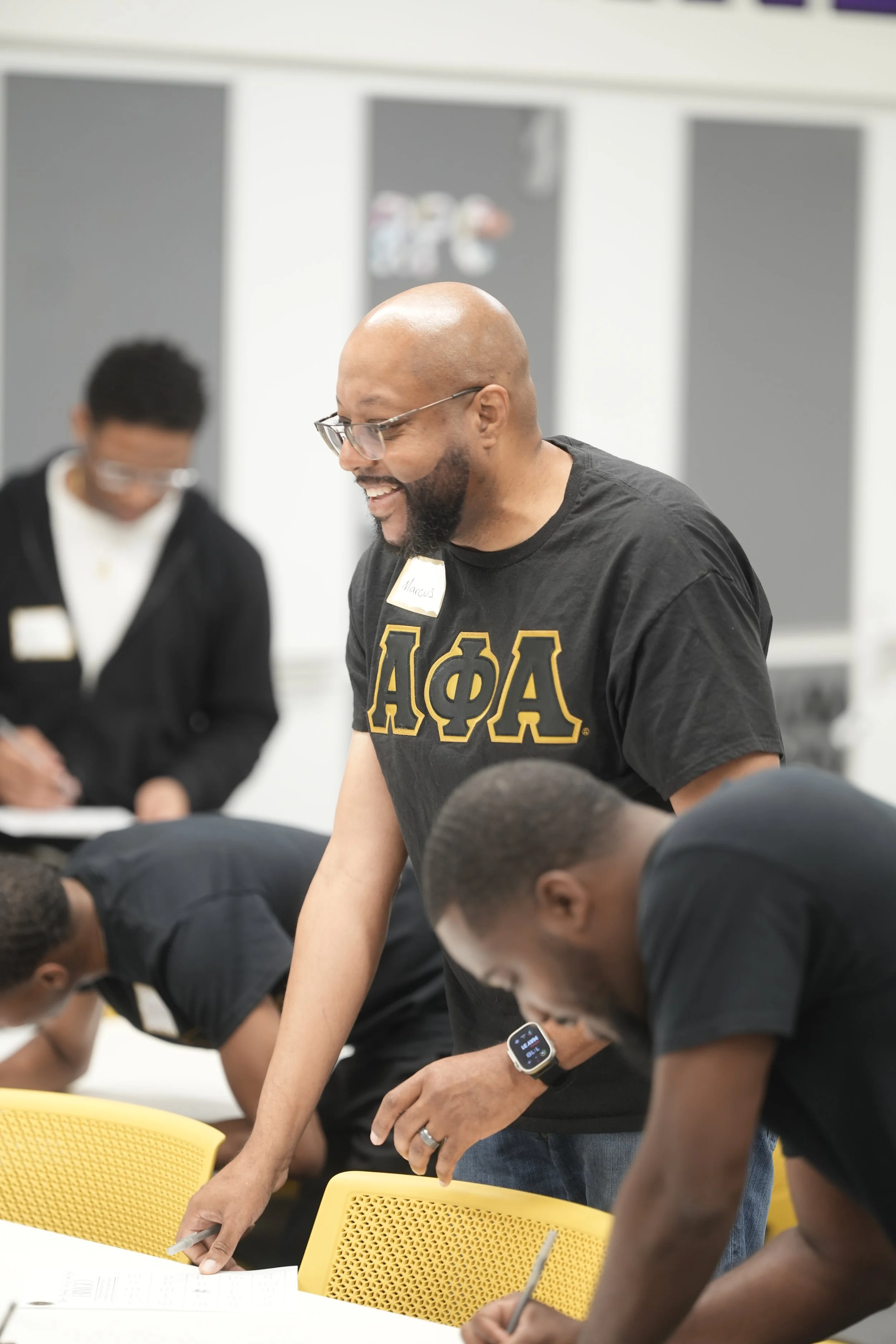 A man wearing glasses and a black T-shirt with Greek letters, smiling and engaged in an activity with others in a classroom setting.