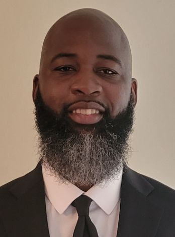 A man with a bald head and a beard wearing a black suit, white shirt, and black tie