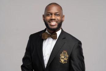 A smiling man in a black suit with a crest on the chest, wearing a white shirt and a patterned bow tie, standing against a plain gray background.