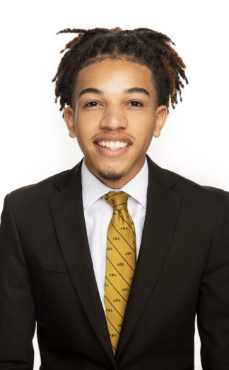 Young man with dreadlocks wearing a black suit, white shirt, and yellow tie, smiling against a white background.