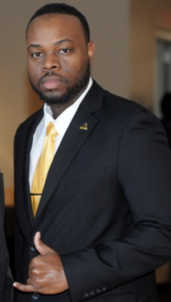 A man in a black suit with a white shirt and yellow tie, standing indoors with a serious expression.