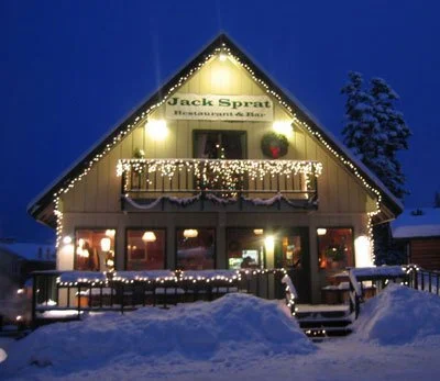 A cozy, two-story restaurant decorated for Christmas with string lights and a wreath, illuminated at night, surrounded by snow.