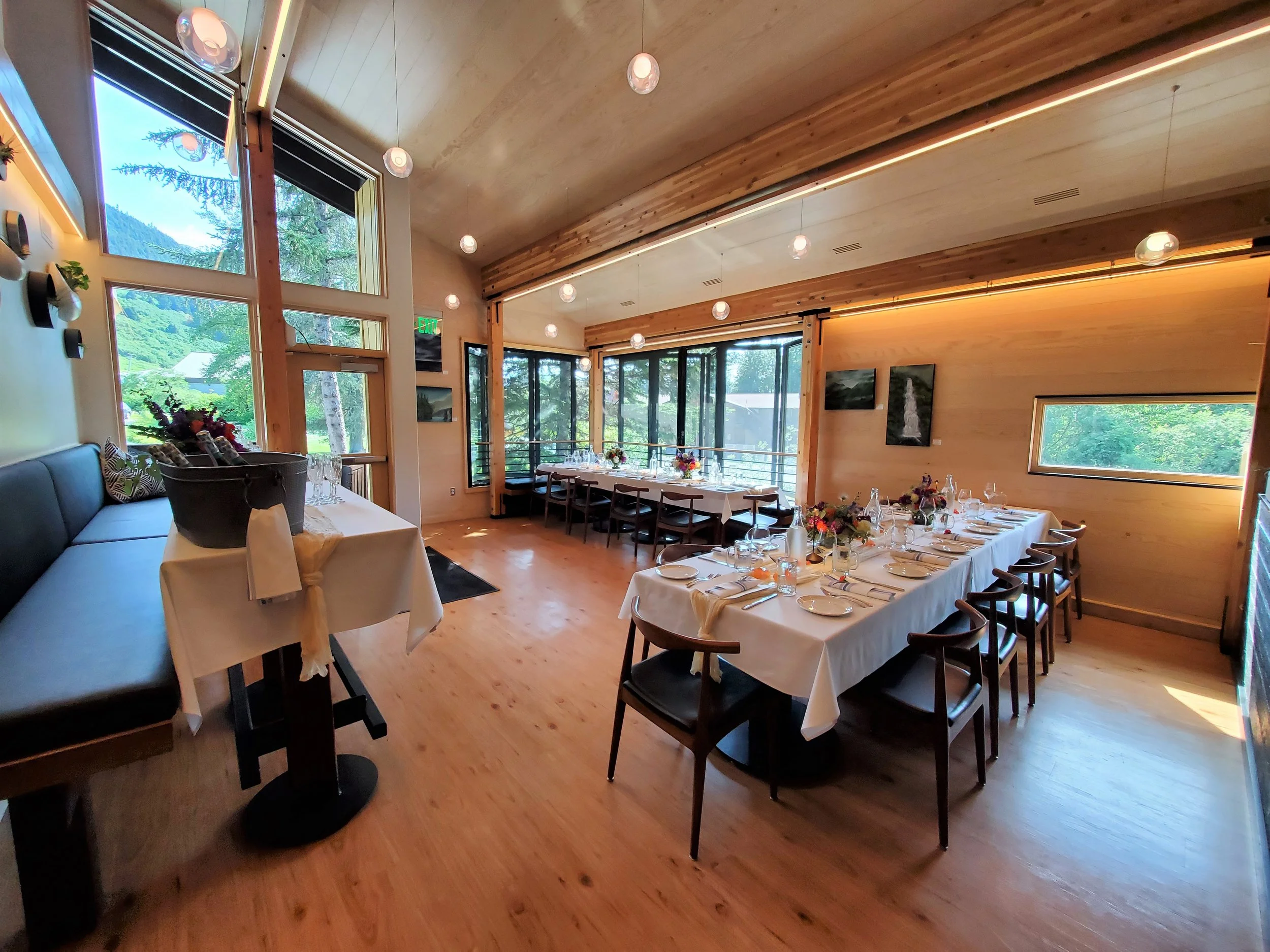 A decorated dining room with large windows overlooking a green landscape, a long table set with white tablecloths, flowers, and tableware, warm wooden interior, and modern lighting.