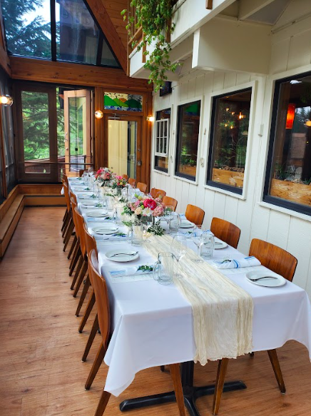 A long dining table set for a meal in a bright, wood-paneled room with large windows. The table is covered with a white tablecloth, decorated with floral arrangements, and set with plates, glasses, and napkins. Wooden chairs line both sides of the ta