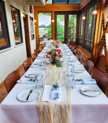 Long dining table set for a meal with white tablecloths, floral centerpieces, and place settings with plates, glasses, and napkins, in a sunlit enclosed porch with wooden walls and large windows.