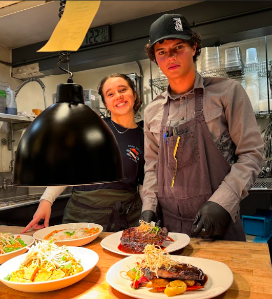 Two cooks behind a kitchen counter, displaying plated dishes including grilled meat and salads. A brother and sister working hard together in a family owned restaurant.