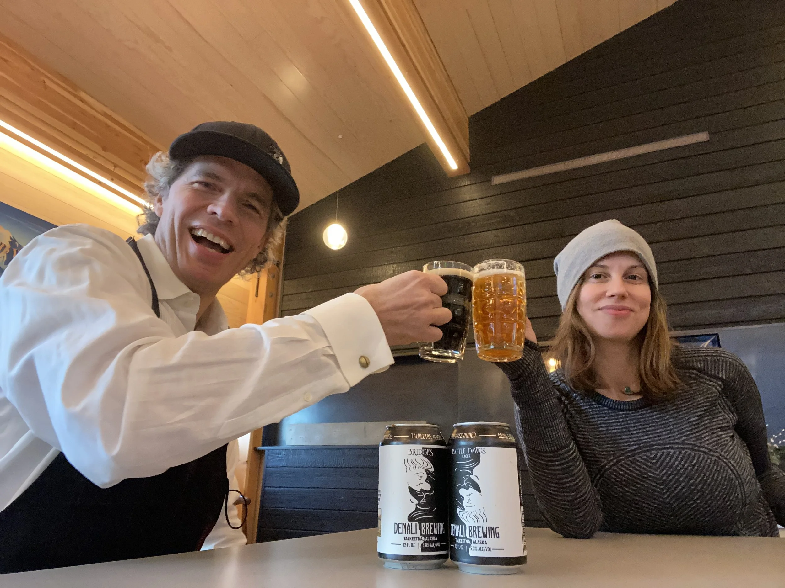 Dynamic duo restauranteurs husband and wife team, Frans and Jen smiling and desperatly in love. Holding beer mugs filled with Denali Brewing beer jugs are on the table in front of them.